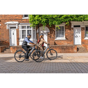 Man and woman riding Dawes Spire 2.0 Cues Crossbar electric hybrid bikes along a residential street with brick houses.