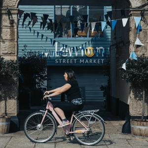 Woman riding the pink Emu Classic Step Through electric bike through the stone entrance of Lemon Street Market.