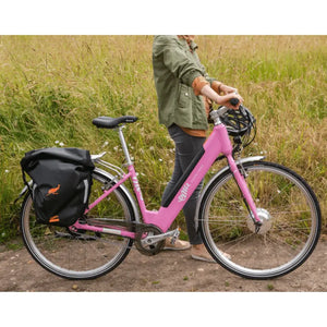 Woman standing with the pink Emu Classic electric bike equipped with a black pannier bag in the countryside.