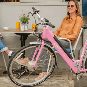 Woman smiling and relaxing at an outdoor cafe table with her pink Emu Classic electric bike parked nearby.