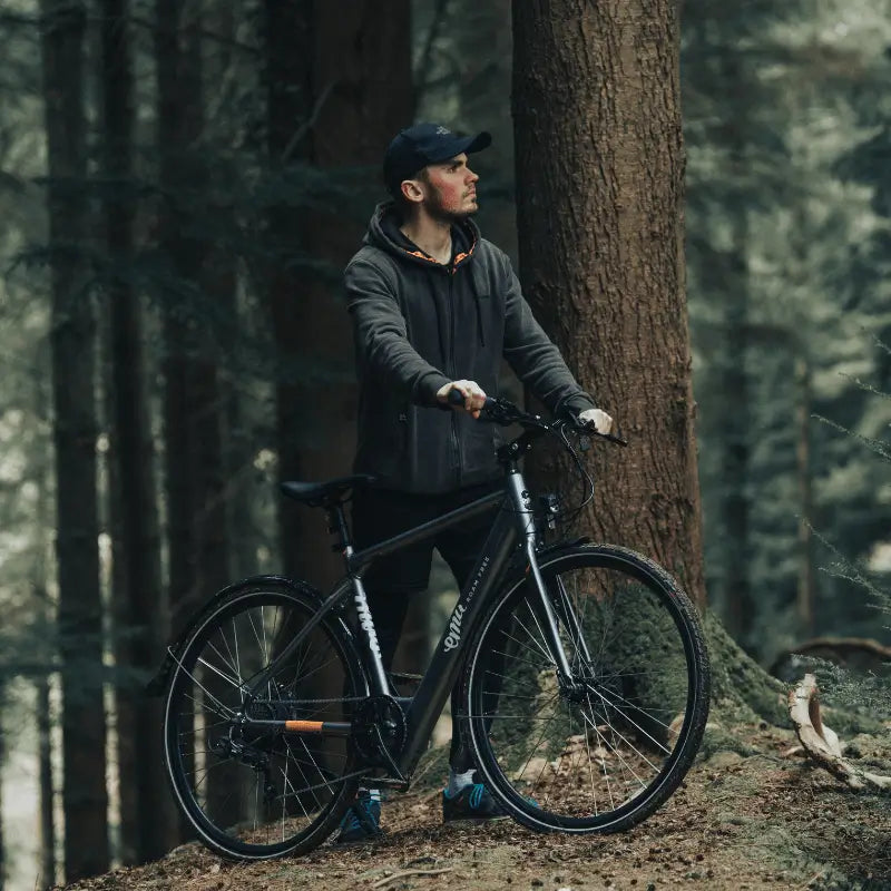 Man standing next to the grey Emu Evo Crossbar electric bike in a dense pine forest setting.