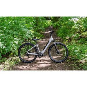 Falcon Horizon Low Step electric hybrid bike in silver parked on a forest trail, surrounded by lush green foliage.