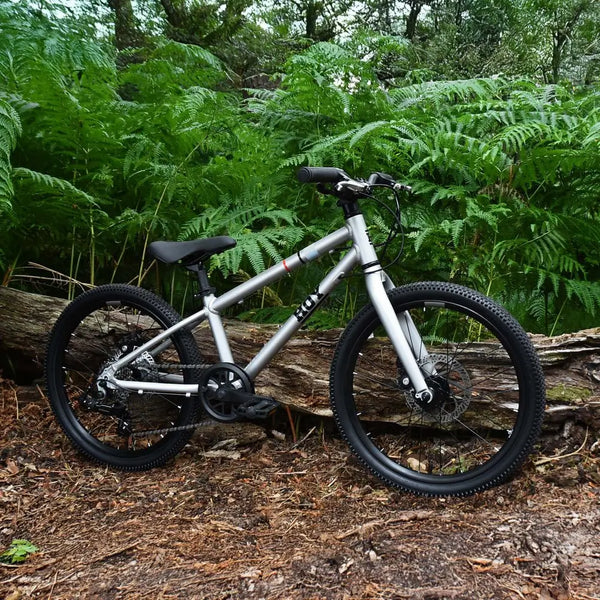 The HOY Pentland black kids' bike in silver leaning on a log in a forest setting, demonstrating its trail capability.