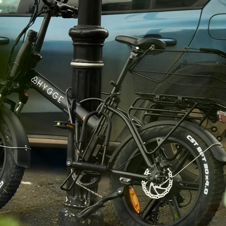 Black Hygge folding electric bike secured to a street post using a heavy-duty Hygge D-lock and cable combination for theft prevention.