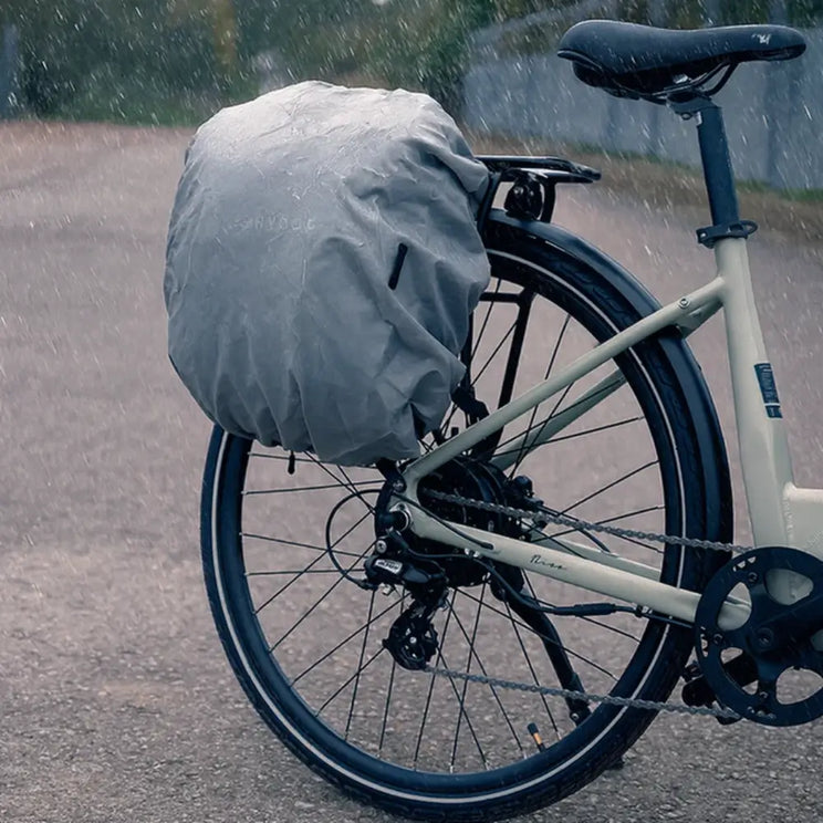 A close-up of a Hygge pannier bag covered by its grey waterproof rain cover attached to a bike rack, protecting contents during a rainy ride.