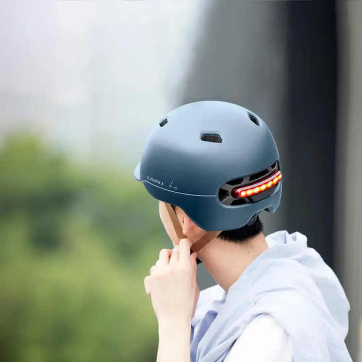 Back view of a person adjusting a blue Livall C20 smart cycle helmet with LED lights, outdoors with a blurred city background.