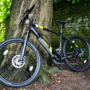 Black Mark2 Scrambler lightweight hardtail e-bike leaning against a tree in a woodland setting.