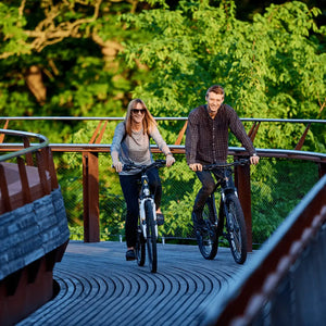 Couple enjoying a leisure ride on Mark2 X-Cross 520 hybrid electric bikes across a wooden bridge.