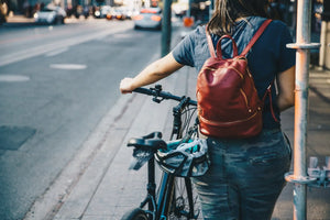 Person with a red backpack standing next to a bicycle on a city street.