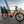 An orange Revom Multi C2 cargo e-bike with child seats and front basket, parked by a river in a city with a bridge in the background at sunset.