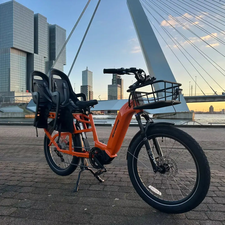 An orange Revom Multi C2 cargo e-bike with two seats and front basket, parked by a river in a city with a bridge in the background at sunset.