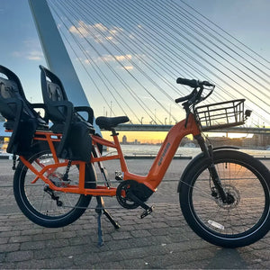 An orange Revom Multi C2 cargo e-bike with two seats and front basket, parked by a river in a city with a bridge in the background at sunset.