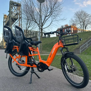 An orange Revom Multi C2 cargo e-bike with two seats and front basket, parked on a paved path in an urban area.