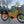 An orange Revom Multi C2 cargo e-bike with two seats and front basket, parked on a paved path in an urban area.
